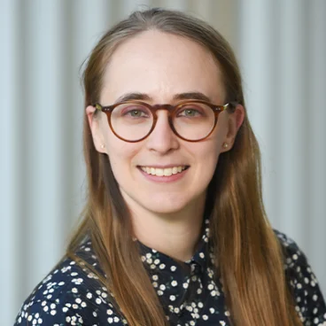 A young woman with long brown hair and round glasses smiles at the camera. She is wearing a dark, patterned blouse and is posed in front of a background with vertical light-colored lines.