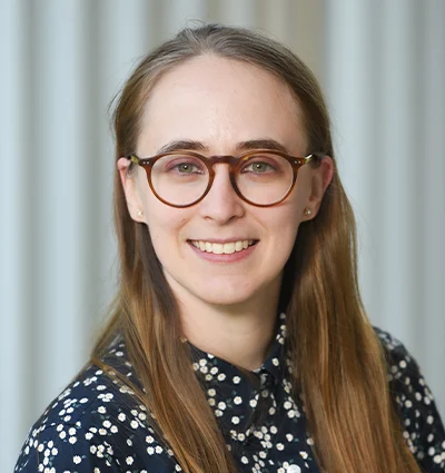 A young woman with long brown hair and round glasses smiles at the camera. She is wearing a dark, patterned blouse and is posed in front of a background with vertical light-colored lines.