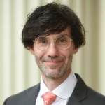 A man with wavy dark hair, glasses, and a short beard is wearing a dark suit, white shirt, and orange patterned tie. He is smiling slightly and standing in front of a light-colored, blurred background.