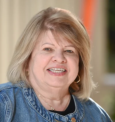 A smiling older woman with shoulder-length blonde hair, wearing a denim jacket and gold hoop earrings, stands in front of a softly blurred background.