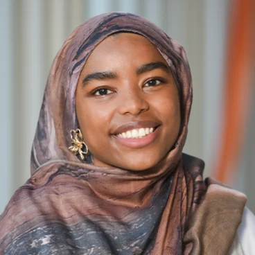 A young woman wearing a patterned brown hijab and a gold butterfly earring smiles warmly in front of a softly blurred background with vertical lines.