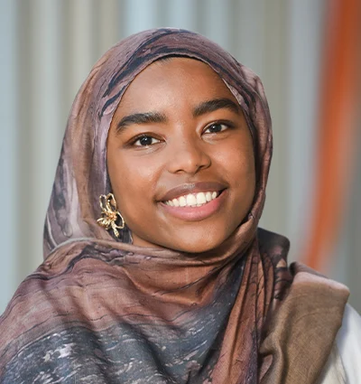 A young woman wearing a patterned brown hijab and a gold butterfly earring smiles warmly in front of a softly blurred background with vertical lines.