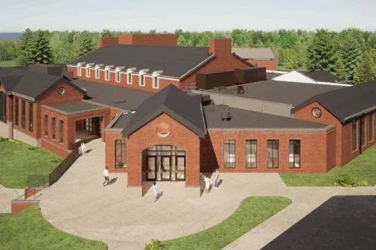 Aerial view of a large, modern brick building with multiple pitched roofs, large windows, and people walking on pathways surrounded by green grass and trees—an ideal location for conference facility rental.