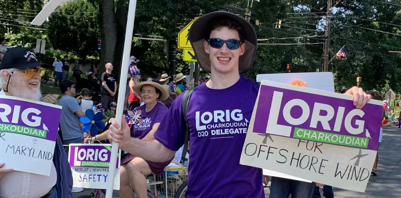 A smiling young man in sunglasses and a wide-brim hat holds a sign supporting Lorig Charkoudian for delegate and offshore wind at an outdoor event. Others nearby also wear purple campaign shirts and hold signs.