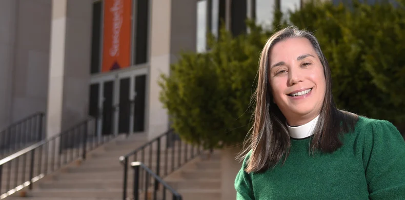 A smiling woman in a green top and white clerical collar stands outside in front of steps and a building with glass doors and a red vertical banner. Green foliage is visible behind her.