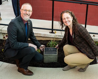 A man and a woman kneel next to a plaque mounted on a stone in a garden bed, both smiling at the camera in front of a red building.
