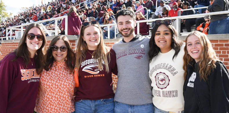 Five smiling people stand together outdoors, wearing 体育买球 University and River Hawks shirts, with a crowded stadium and brick wall in the background. It appears to be a sunny day at a sporting event.