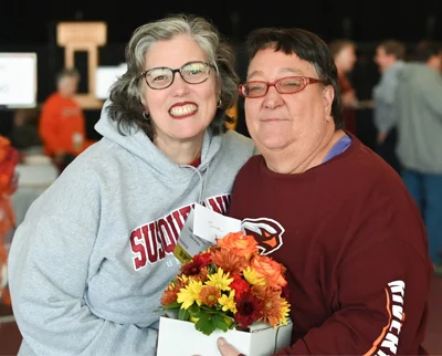Two smiling women stand close together indoors. One holds a bouquet of colorful flowers. Both wear glasses and casual sweatshirts, and appear happy and friendly.