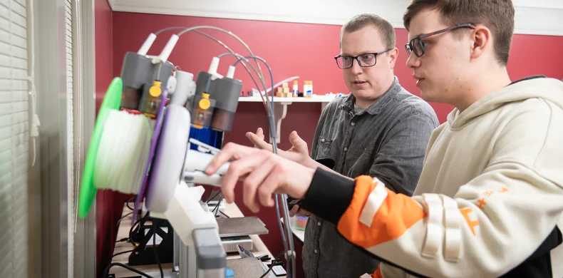 Two people work together at a machine with spools of filament, possibly a 3D printer, in a room with red walls. One person points at the machine while the other adjusts its controls.