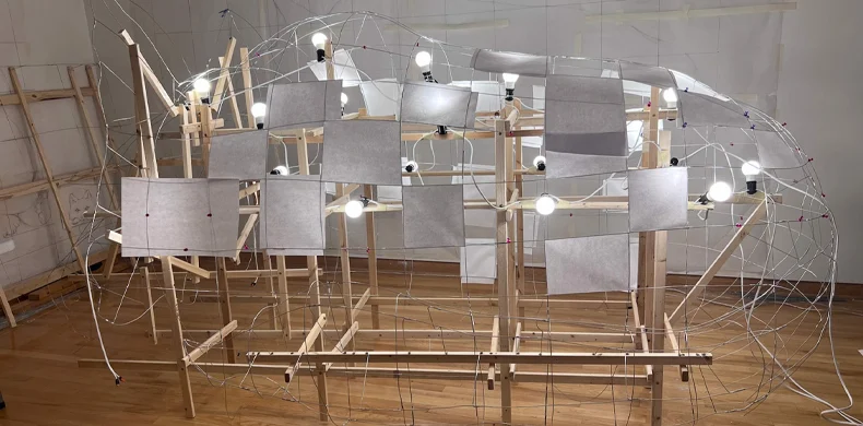 A large, abstract structure made of wooden beams, wire frames, and hanging white panels is displayed indoors. Several light bulbs are attached throughout the installation, illuminating the scene.
