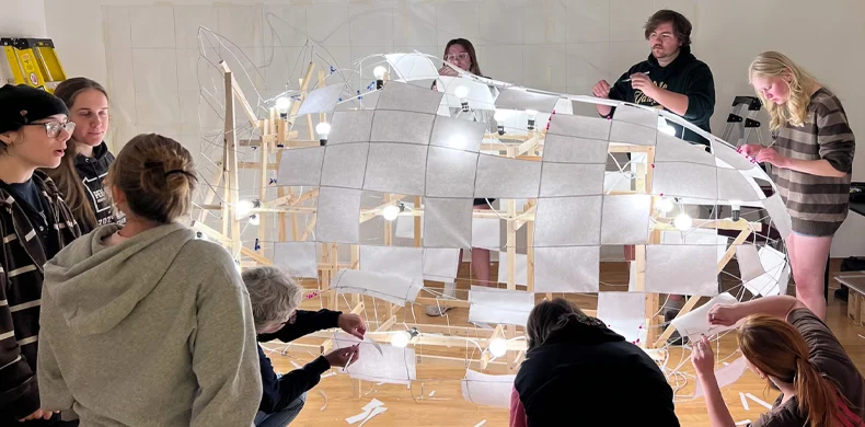 A group of people work together to construct a large, illuminated art installation using wooden frames, wire, and white panels in a brightly lit indoor space.