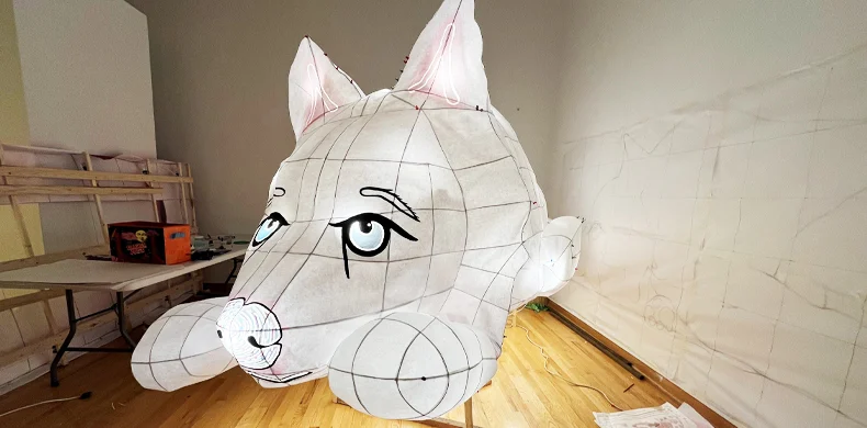 A large, unfinished animal lantern shaped like a cat’s head, made of white translucent material with black facial features, stands on a wooden floor in a sparsely furnished room.