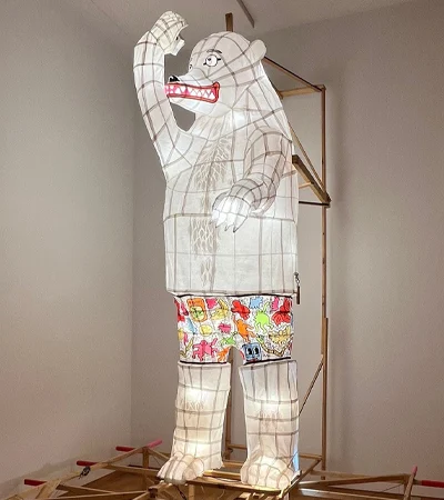 A large, illuminated bear-shaped lantern with a grid-like frame, wearing colorful cartoon-printed shorts, stands on a wooden structure indoors, raising one paw and showing its teeth.