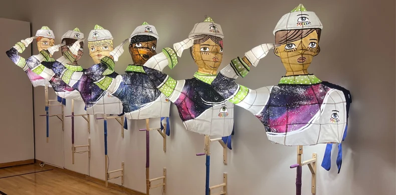Six large, colorful puppet figures with illuminated faces and arms raised in a salute, wearing white hats and jackets, are mounted on wooden stands against a gallery wall.