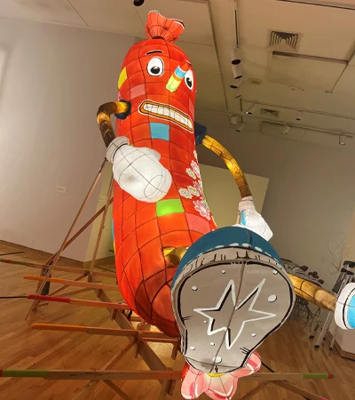 A large, colorful, cartoonish sausage sculpture with a patchwork pattern, exaggerated facial features, and oversized shoes sits on a wooden frame in a gallery space with wooden floors and white walls.