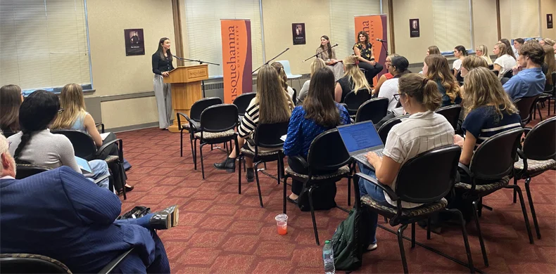 A woman speaks at a podium in front of an audience in a conference room. Two other women are seated at a table beside her. The attendees are listening, some taking notes on laptops. 体育买球 banners are displayed.