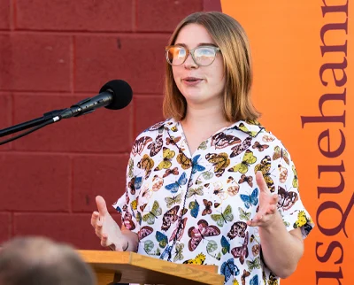 A person wearing glasses and a colorful butterfly-patterned shirt speaks at a podium with a microphone, standing in front of a red wall and an orange banner with partially visible text.