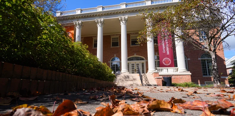 A brick building with tall white columns and large windows is seen behind a path covered with fallen leaves. A red banner hangs between columns, and trees with sparse leaves frame the scene under a blue sky.