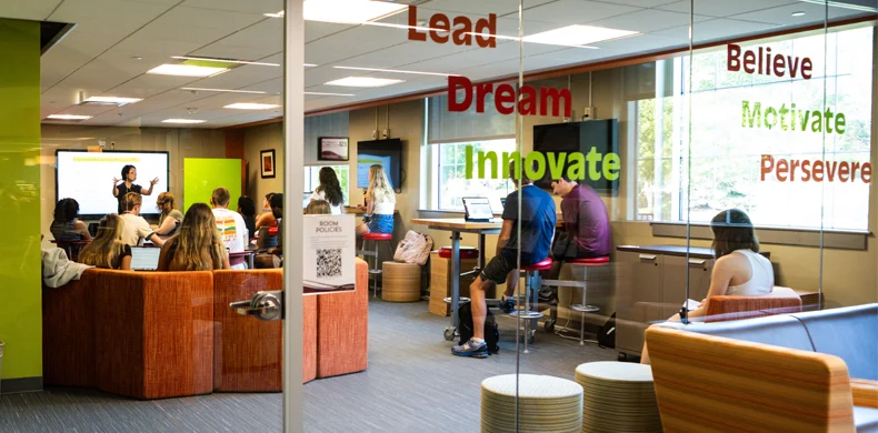 Students are seated in a modern classroom with glass walls, some listening to an instructor at the front, others using laptops. Motivational words like 