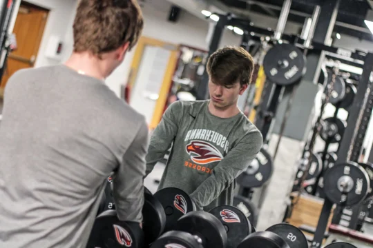 A young man in a long-sleeve shirt selects dumbbells from a rack in a gym, looking down with focus. His reflection is visible in a mirror behind the weights.