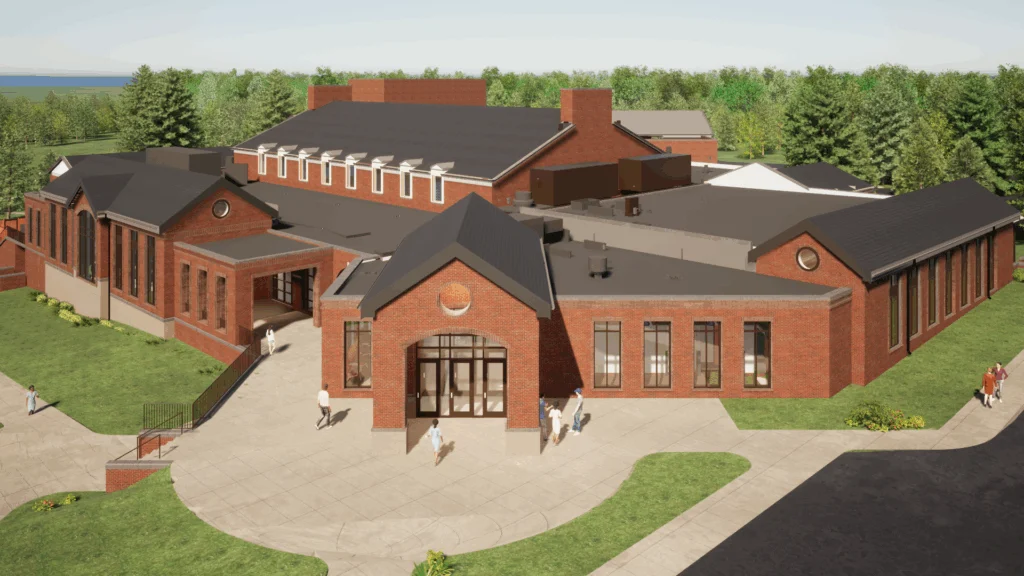 Aerial view of a large brick building with multiple peaked roofs, large windows, and pathways highlights Campus Transformations. People walk near the main entrance, surrounded by trees and greenery in the background.