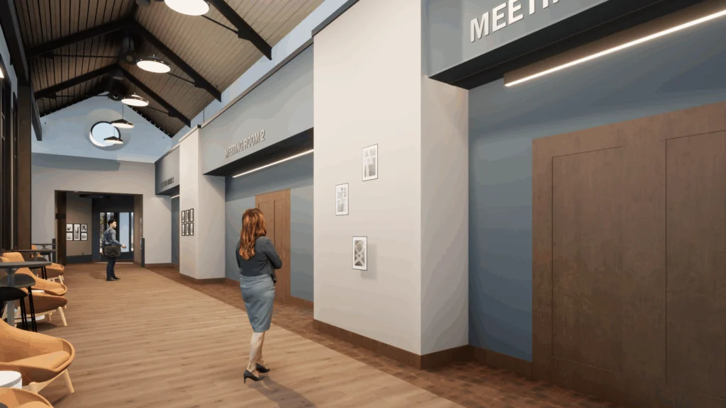 A woman walks down a modern hallway with wood floors, gray walls, and high ceilings—part of recent Campus Transformations. Meeting rooms and offices line the corridor as framed pictures adorn the walls. Another person stands near the entrance.