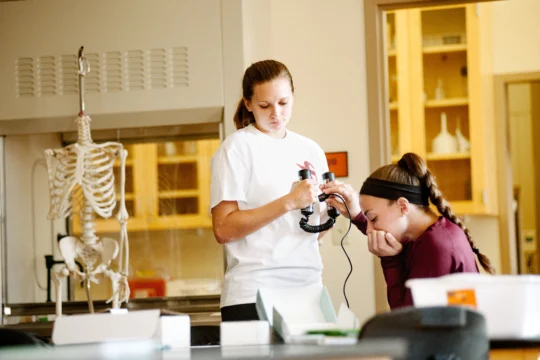Two students in a science classroom; one uses an otoscope to examine the other’s ear. A human skeleton model and lab equipment are visible in the background.