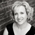 Black and white photo of a smiling woman with short, curly blonde hair, wearing a dark top, leaning against a brick wall.
