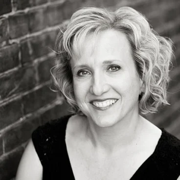 Black and white photo of a smiling woman with short, curly blonde hair, wearing a dark top, leaning against a brick wall.