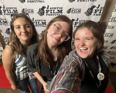 Three young women smile for a selfie in front of an 