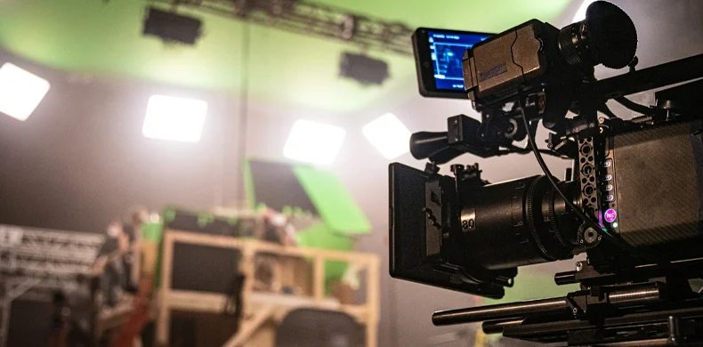 A professional video camera is set up on a film set with bright lights, scaffolding, and a green screen in the background, indicating a movie or television production in progress.