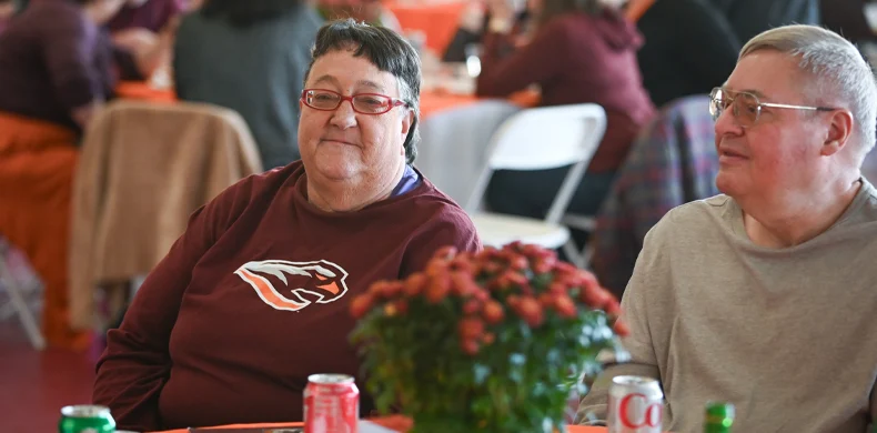 Two people sit at a table with drinks and a potted plant, smiling. The woman wears glasses and a maroon shirt with a beaver logo. Other people are seen in the blurred background at a gathering.