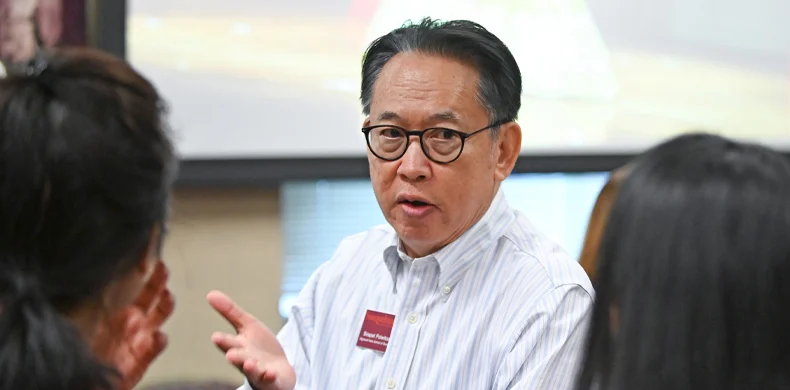 A man wearing glasses and a striped shirt speaks to a small group of people in an indoor setting. He gestures with one hand, and a projector screen is visible in the background.