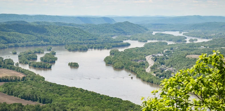 A wide river curves through a lush, green landscape with rolling hills in the background. Trees line the riverbanks, and a road runs alongside the river. The sky is partly cloudy.