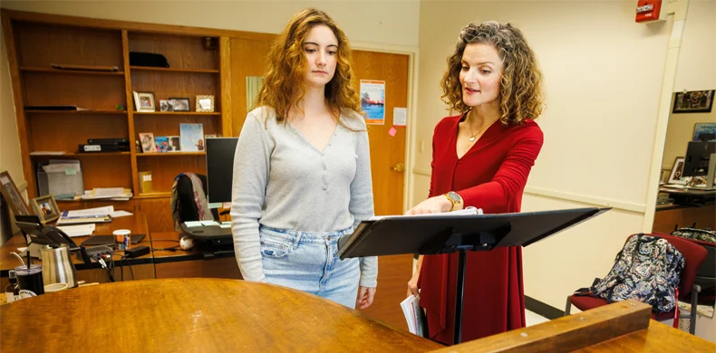 Two women stand beside a piano in an office. One, in a red dress, points to a music stand while the other, in a gray top and jeans, listens attentively. Shelves, a desk, and a backpack are visible in the background.