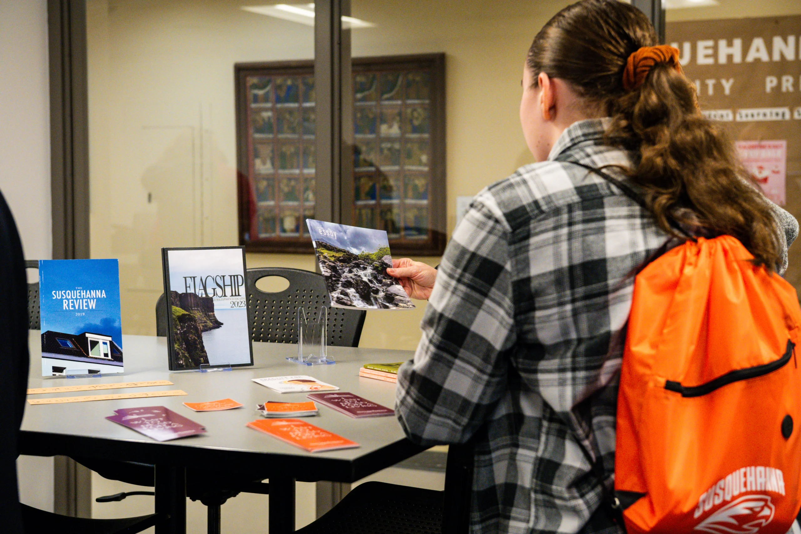 A person with a plaid shirt and orange backpack looks at a magazine at a table displaying various books and brochures in a bright indoor setting.