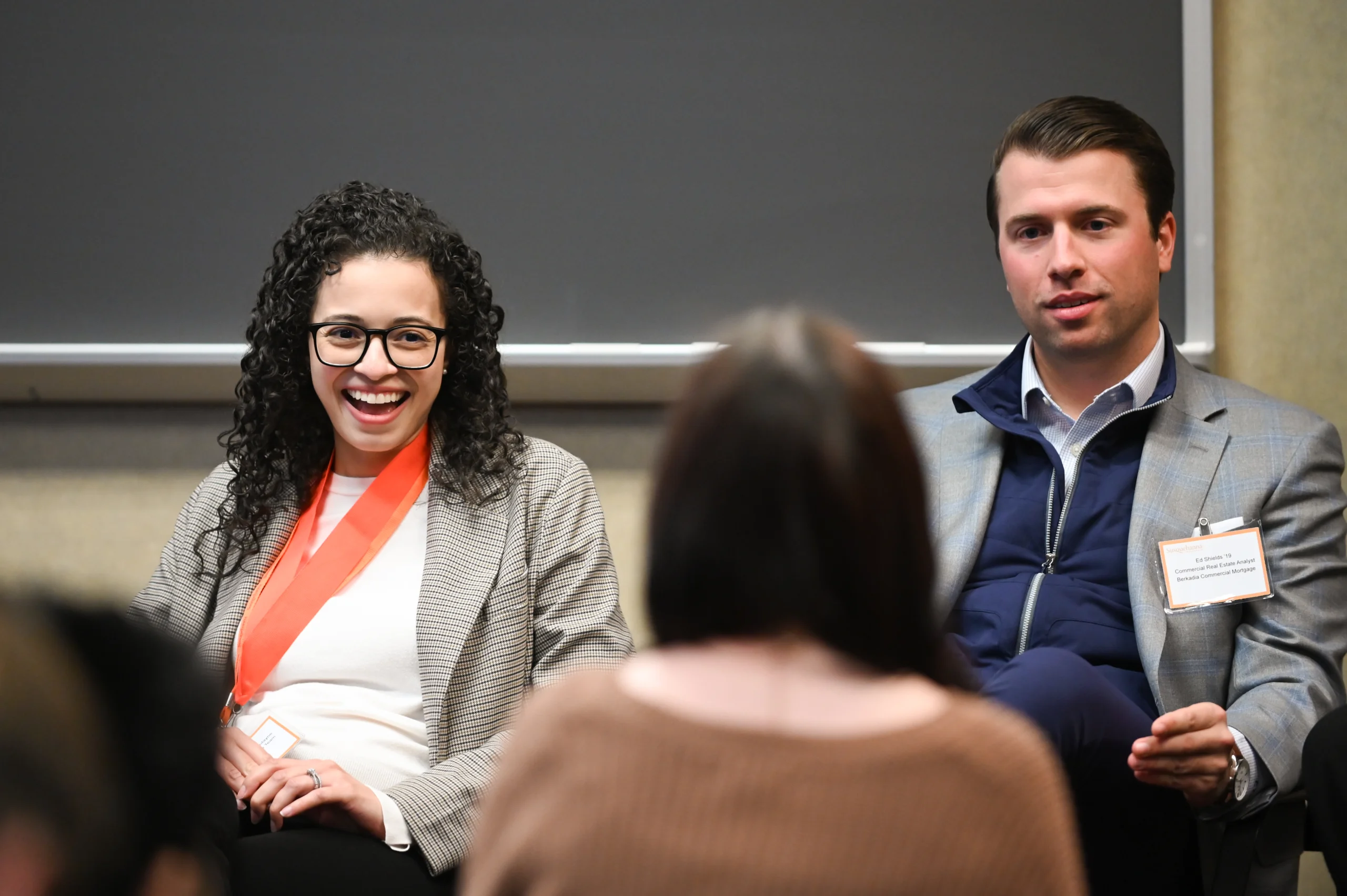 Two people sit side by side in front of a chalkboard, engaged in conversation with someone facing them. The woman smiles brightly, wearing glasses and a suit, while the man sits calmly in a blazer with a name tag.