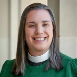 A smiling woman with straight, shoulder-length brown and gray hair wears a green sweater and a white clerical collar, standing in front of a light-colored pillar.