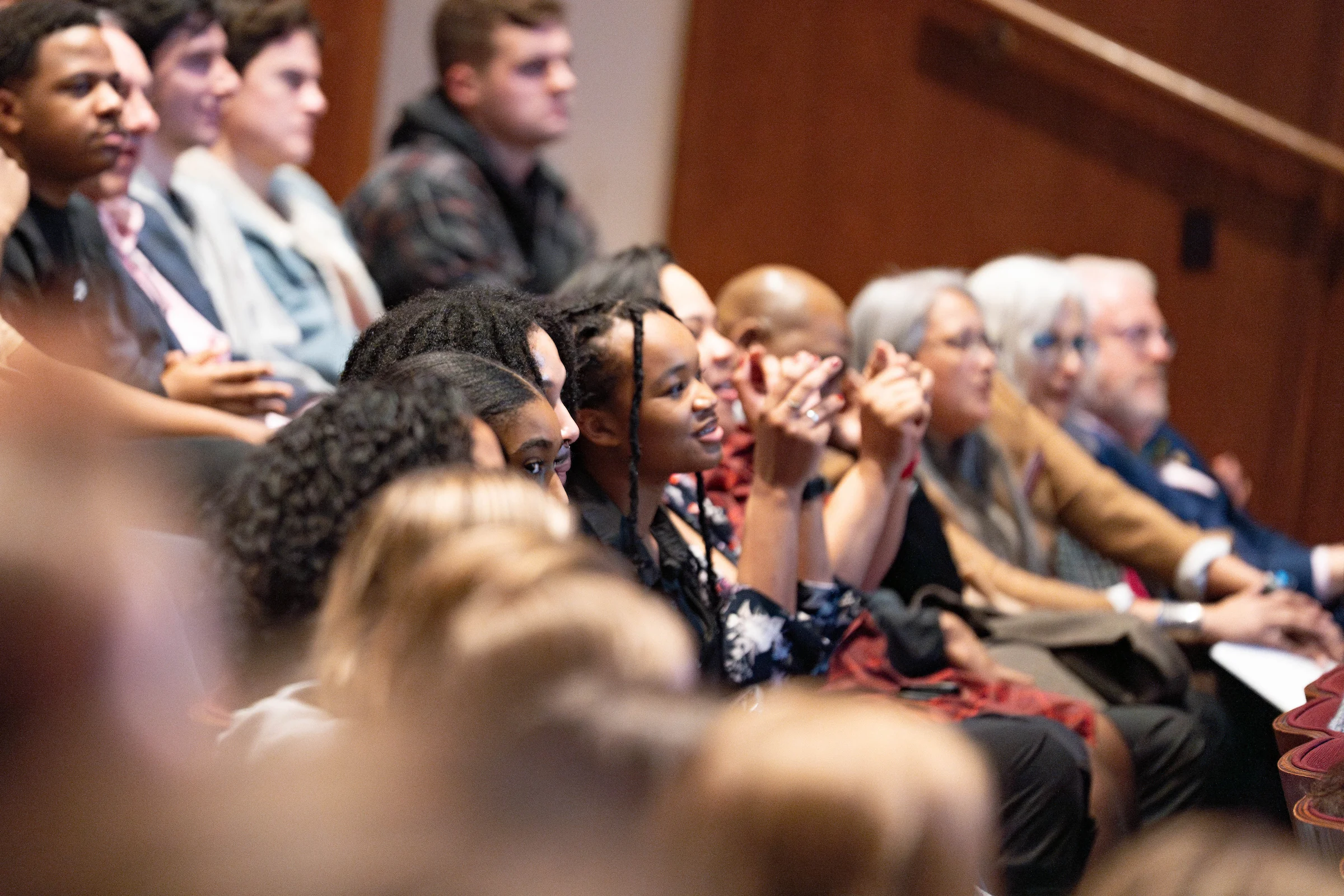 A diverse group of people sit closely together in an auditorium, attentively watching an event. Some are smiling and clapping, while others look focused. The lighting is warm and the mood appears engaged and positive.