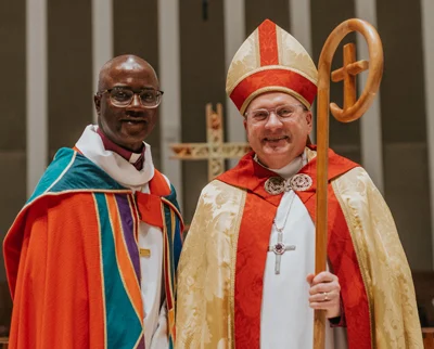 Two clergy members in colorful ceremonial robes stand smiling side by side. One wears a gold and red miter and holds a staff; the other wears glasses and a multicolored robe. A cross is visible in the background.
