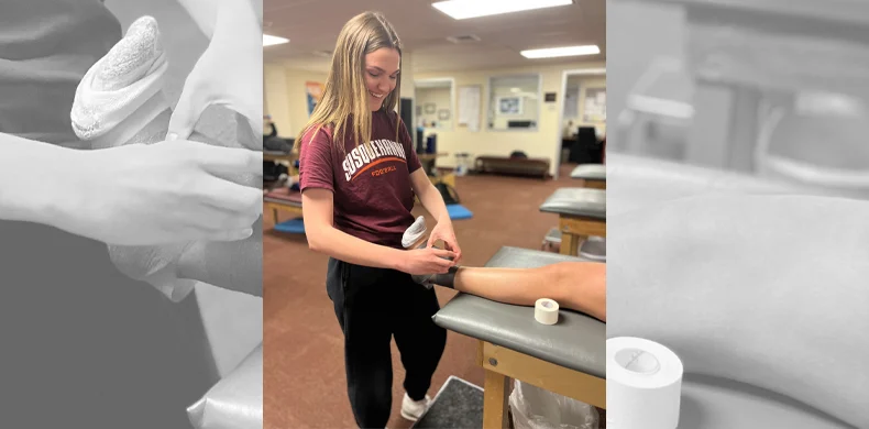 A young woman smiles while wrapping athletic tape around another person’s ankle in a clinic or training room, with rolls of tape and tables visible in the background.