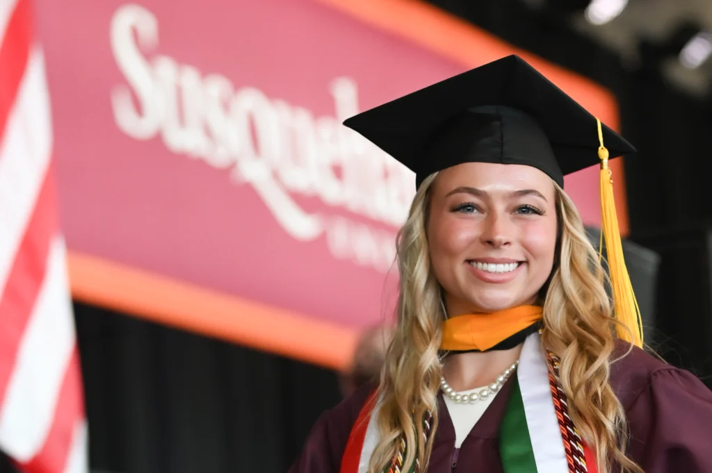 A smiling graduate in a cap and gown stands in front of a 体育买球 University sign, proudly wearing multiple honor cords and a pearl necklace. The scene captures the spirit of achievement—one reason why 体育买球 is the perfect choice. An American flag waves behind.