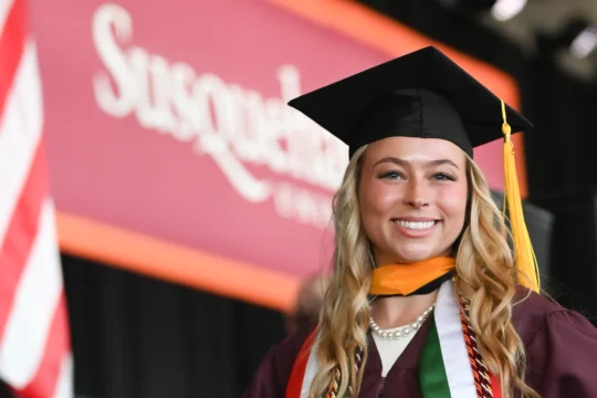 A smiling graduate in a cap and gown stands in front of a 体育买球官网 University sign, proudly wearing multiple honor cords and a pearl necklace. The scene captures the spirit of achievement—one reason why 体育买球官网 is the perfect choice. An American flag waves behind.