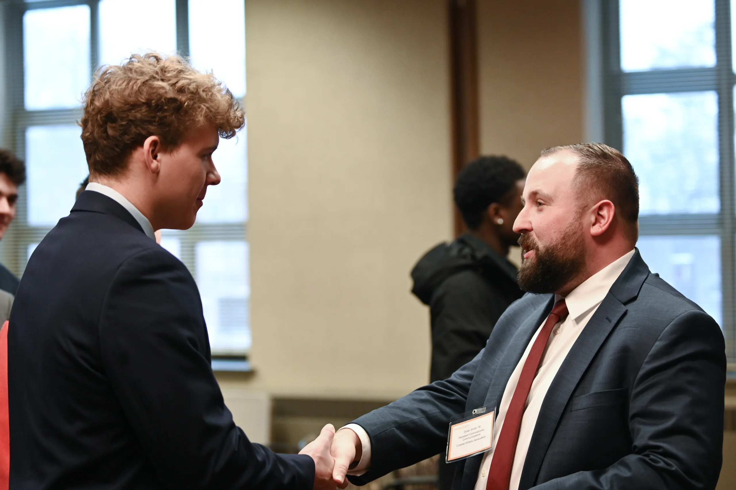 Two men in suits shake hands and smile at an indoor event, with other people and large windows visible in the background.