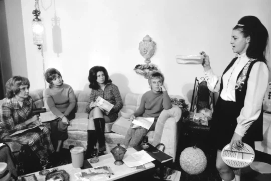 A woman stands and holds up a plastic container, demonstrating it to four seated women in a living room filled with various household items—creating an atmosphere reminiscent of a home product demo at the Lore Degenstein Gallery.