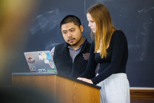 Two students stand at a podium in front of a laptop covered in stickers, focused on the screen as they explore career services resources, with a chalkboard in the background.