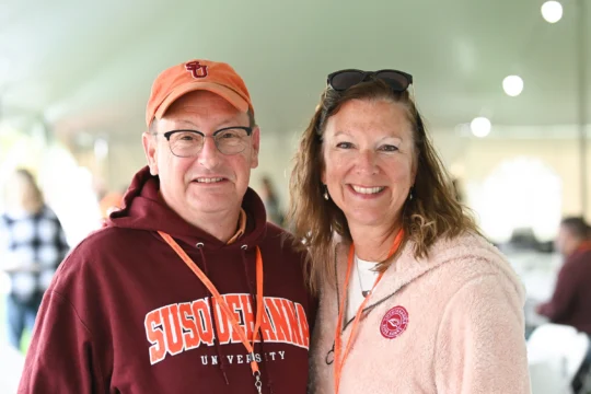 Two adults smiling at the camera under a tent. Wearing orange lanyards, they proudly represent Champions for Growth—he in a “体育买球官网 University” hoodie and orange cap, she in a light pink fleece with sunglasses on her head.