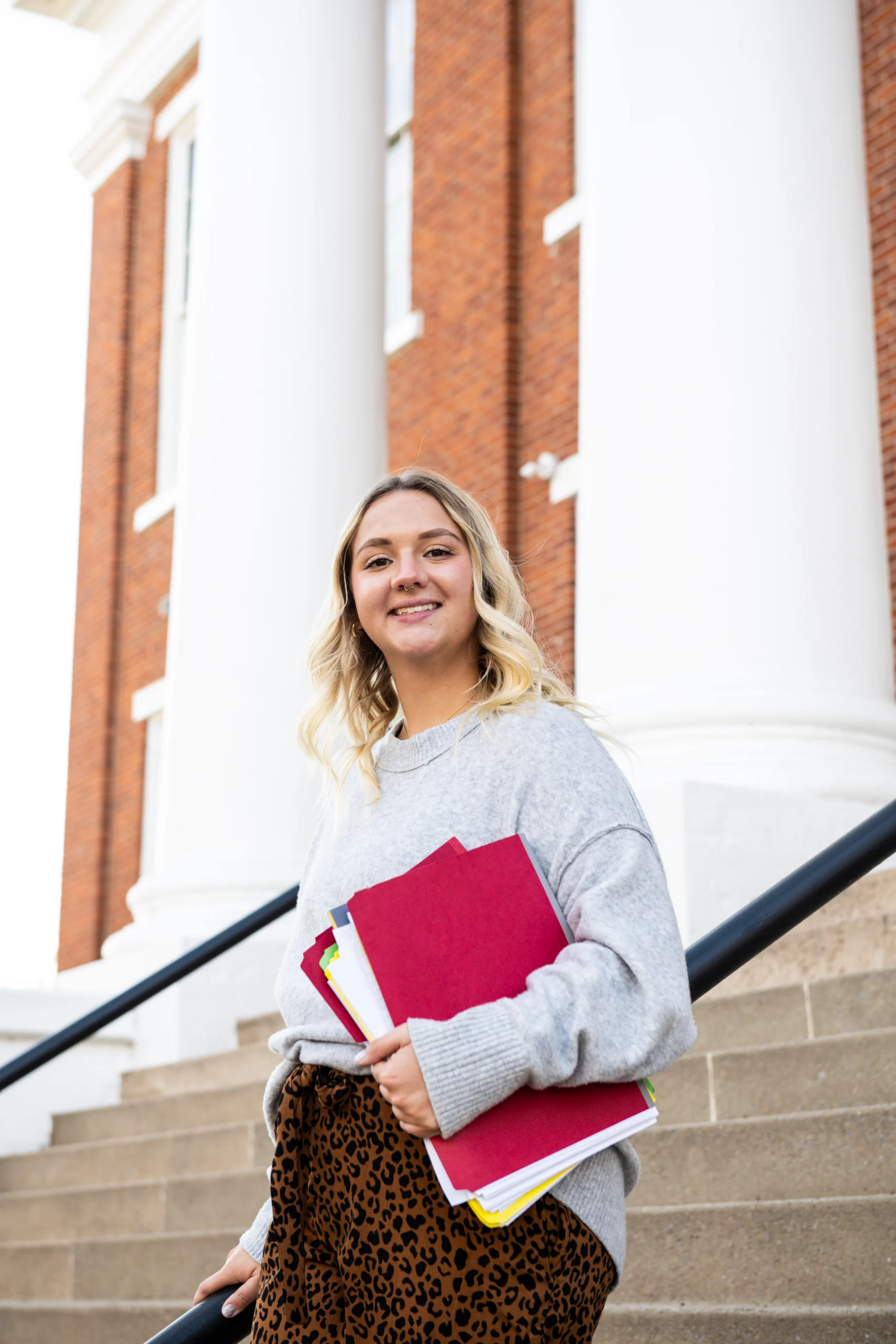 A young woman with blonde hair, wearing a gray sweater and patterned pants, smiles while holding folders—reflecting strong graduate outcomes—as she stands on steps in front of a building with white columns and red brick walls.
