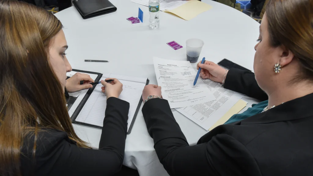 Two women sit at a round table reviewing documents together; one is writing on a notepad while the other holds up a resume and writes on it—an engaging scene of career services in action. The table has papers, a pen, a water bottle, and a cup.