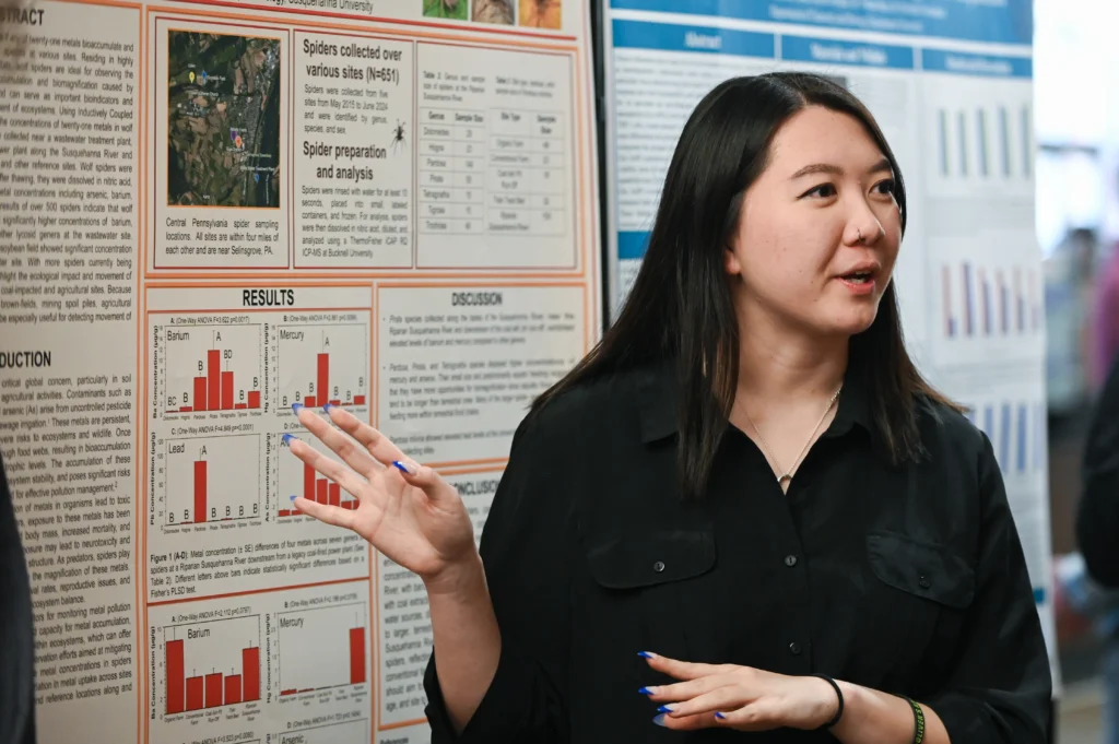 A woman with long dark hair presents research in front of a display board filled with scientific charts, graphs, and text about spiders, highlighting how Funding for Real-World Learning supported her hands-on investigation. She gestures while explaining her findings.